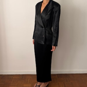 Op de afbeelding: Een zwarte double-breasted blazer met een satijnachtige afwerking, voorzien van revers, lange mouwen en donkere knopen, gecombineerd met een bijpassende zwarte rok tot op de grond en eenvoudige zwarte teenslippers voor een monochrome look.