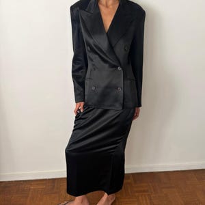 May include: A black satin double-breasted blazer with structured shoulders and notched lapels, paired with a matching black satin floor-length skirt. The blazer features four visible buttons on the front and long sleeves with a smooth, reflective finish.