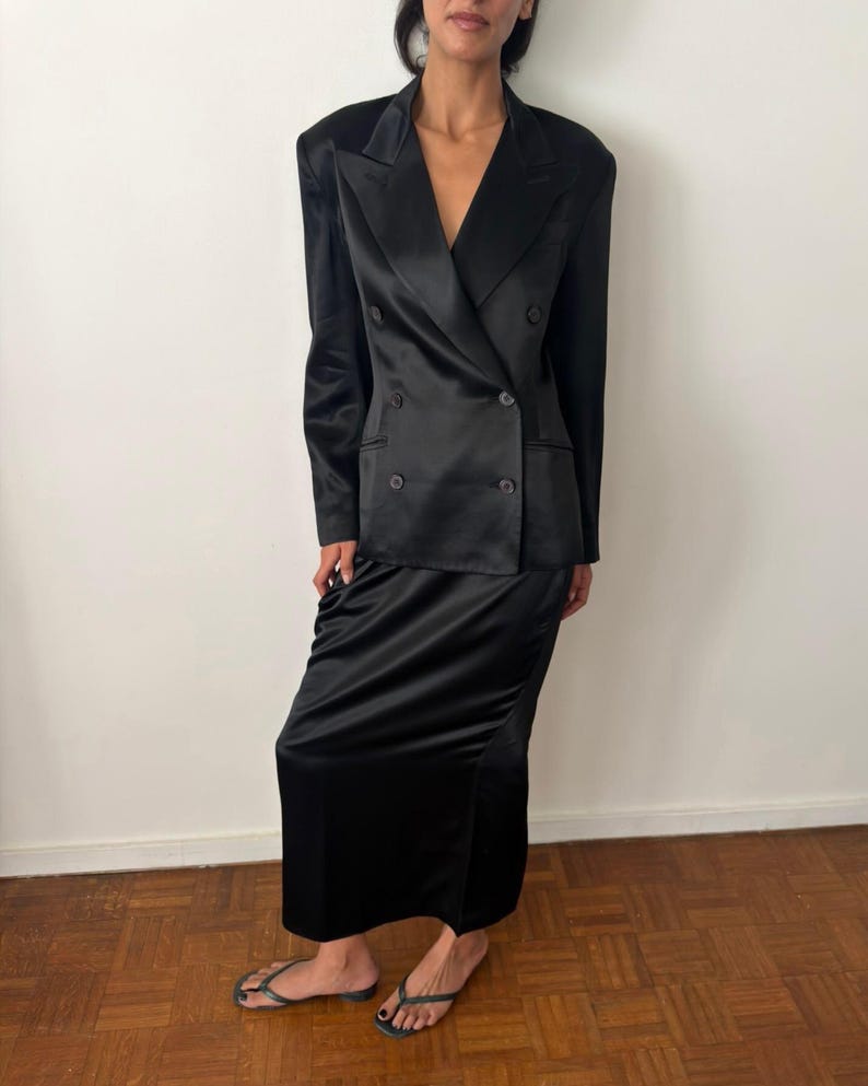 May include: A black satin double-breasted blazer with structured shoulders and notched lapels, paired with a matching black satin floor-length skirt. The blazer features four visible buttons on the front and long sleeves with a smooth, reflective finish.