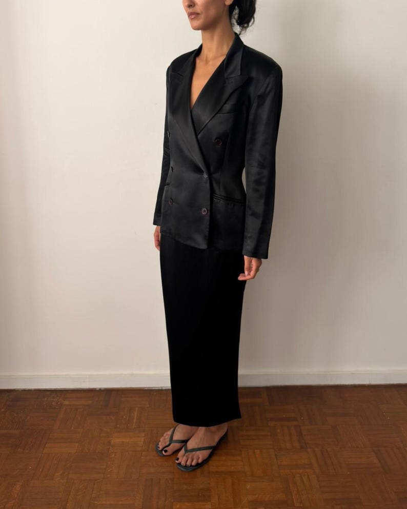 May include: A black double-breasted blazer with a satin-like finish, featuring notched lapels, long sleeves, and dark buttons, paired with a matching black floor-length skirt and simple black thong sandals, creating a monochromatic ensemble.