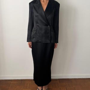 May include: A black double-breasted blazer with a satin finish, featuring wide lapels and four buttons, paired with a matching black floor-length skirt. The ensemble is constructed from a smooth, dark fabric that reflects light across the surface.