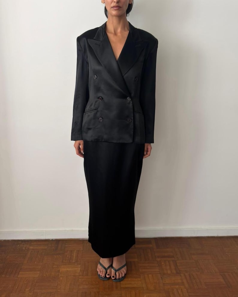 May include: A black double-breasted blazer with a satin finish, featuring wide lapels and four buttons, paired with a matching black floor-length skirt. The ensemble is constructed from a smooth, dark fabric that reflects light across the surface.