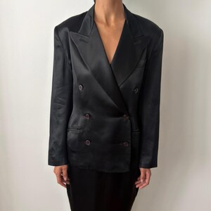 May include: A black double-breasted blazer crafted from a smooth, satin-like fabric. The garment features wide lapels, structured shoulders, and four dark buttons arranged in two columns on the front, along with two side pockets.