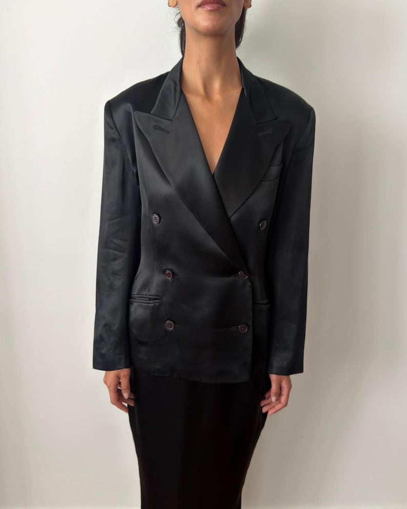 May include: A black double-breasted blazer crafted from a smooth, satin-like fabric. The garment features wide lapels, structured shoulders, and four dark buttons arranged in two columns on the front, along with two side pockets.