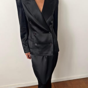 May include: A black satin double-breasted blazer featuring structured shoulders, a notched lapel, and four dark buttons on the front. The blazer is paired with a matching black satin skirt that falls to a midi length, creating a coordinated set.
