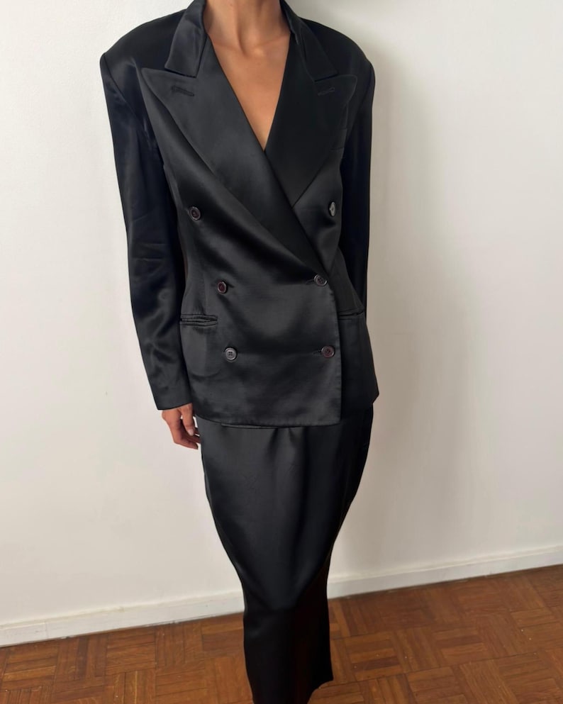 May include: A black satin double-breasted blazer featuring structured shoulders, a notched lapel, and four dark buttons on the front. The blazer is paired with a matching black satin skirt that falls to a midi length, creating a coordinated set.