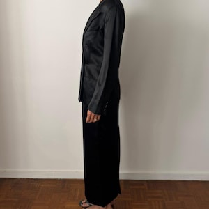 May include: A black satin blazer with a notched lapel, long sleeves, and a buttoned front closure, paired with a matching black floor-length skirt. The ensemble features a smooth, reflective finish and a tailored fit, shown from a side profile view.