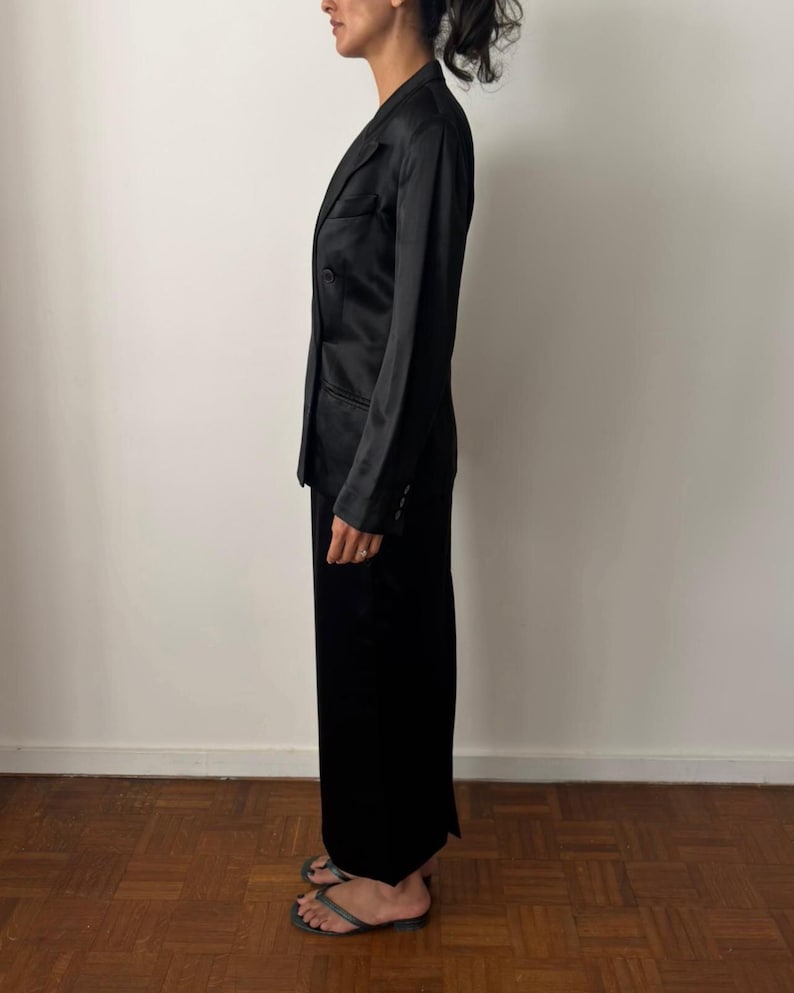 May include: A black satin blazer with a notched lapel, long sleeves, and a buttoned front closure, paired with a matching black floor-length skirt. The ensemble features a smooth, reflective finish and a tailored fit, shown from a side profile view.