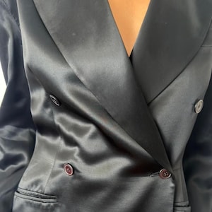 May include: A black double-breasted blazer crafted from a smooth, satin-like fabric with a subtle sheen. The garment features wide lapels, long sleeves, and dark, round buttons arranged in two vertical rows, along with two horizontal welt pockets.