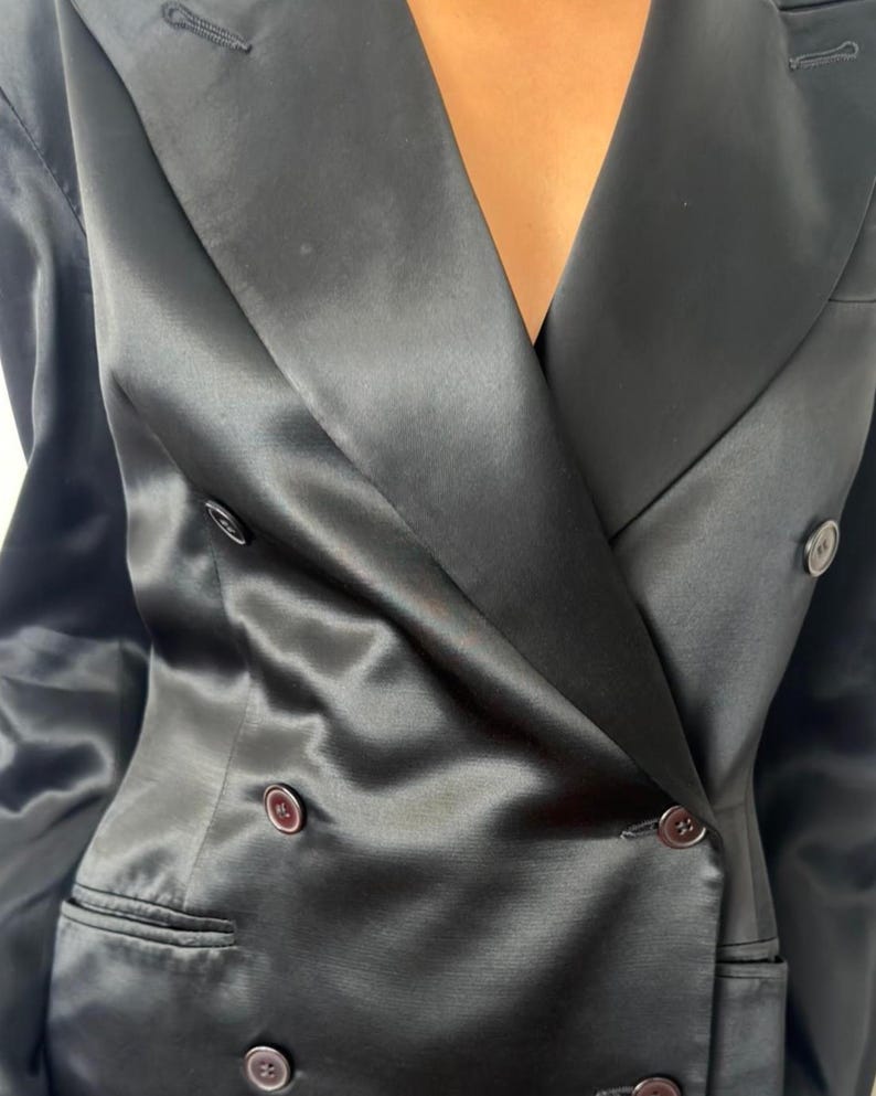 May include: A black double-breasted blazer crafted from a smooth, satin-like fabric with a subtle sheen. The garment features wide lapels, long sleeves, and dark, round buttons arranged in two vertical rows, along with two horizontal welt pockets.