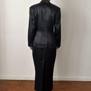 May include: A black satin two-piece set consisting of a long-sleeved blazer with structured shoulders and a matching floor-length skirt. The blazer features a tailored fit with a buttoned front, while the skirt has a straight silhouette.