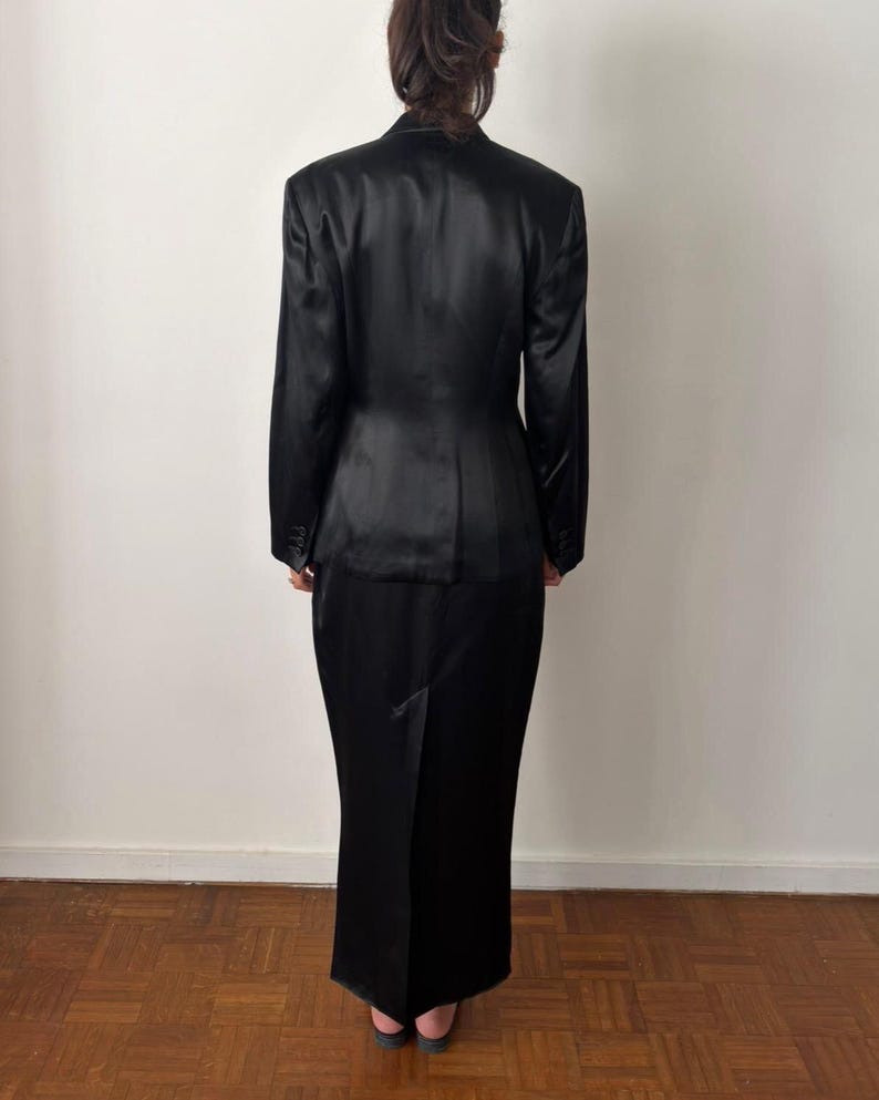May include: A black satin two-piece set consisting of a long-sleeved blazer with structured shoulders and a matching floor-length skirt. The blazer features a tailored fit with a buttoned front, while the skirt has a straight silhouette.
