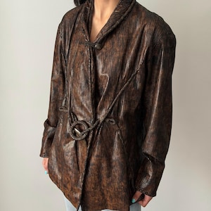 May include: A brown and black textured jacket with a unique design. The jacket features a wrap-style front with a rope tie and a decorative button closure. The long-sleeved jacket has a metallic sheen, adding a touch of glamour to the garment.