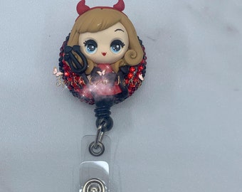 Pode incluir: Carretel de crachá de brilho vermelho e preto com uma imagem de desenho animado de uma menina com chifres vermelhos e um garfo. A menina está vestindo um vestido vermelho e tem cabelo loiro.
