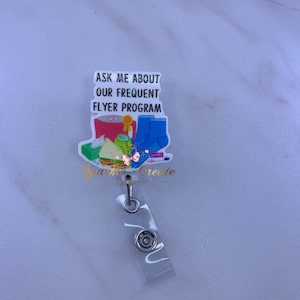 May include: White badge reel with a colorful graphic that says "Ask me about our frequent flyer program". The graphic includes a cup of coffee, a sandwich, a roll of toilet paper, a cucumber, a pair of socks, and a tube of toothpaste.