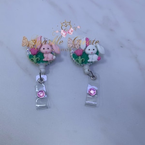 May include: Two white and pink bunny themed retractable badge holders with a green and pink floral design, pink rhinestones, and a butterfly charm. The badge holders have a clear plastic strap with a pink rhinestone button.