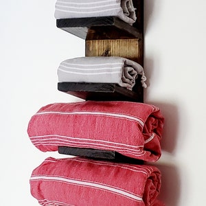 May include: A rustic wooden towel rack with three shelves. The shelves hold rolled towels in gray and red with white stripes. The wood has a dark stain, creating a contrast with the light-colored towels.