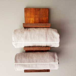 May include: A wooden towel rack with a dark brown finish, holding two rolled gray towels with white trim. The rack is mounted on a light gray wall, showcasing a minimalist design.