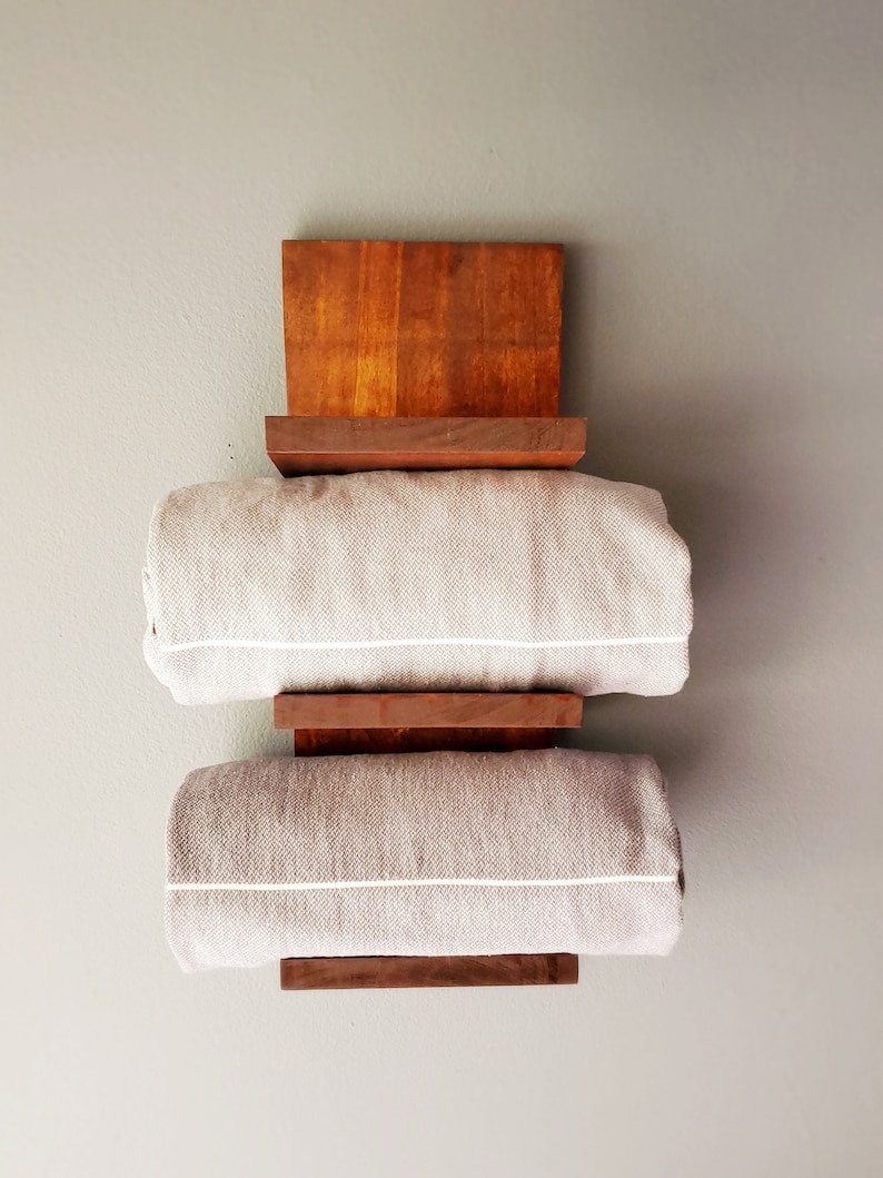 May include: A wooden towel rack with a dark brown finish, holding two rolled gray towels with white trim. The rack is mounted on a light gray wall, showcasing a minimalist design.