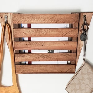 May include: A wooden mail organizer with a slatted design, stained in a warm brown hue. The organizer is mounted on a white wall and features two metal hooks. A yellow bag, keys, and a small purse hang from the hooks.