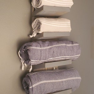 May include: A wooden wall-mounted towel rack with three shelves, each holding a rolled towel. The towels are beige and blue, with white stripes. The rack is painted gray and mounted on a gray wall.