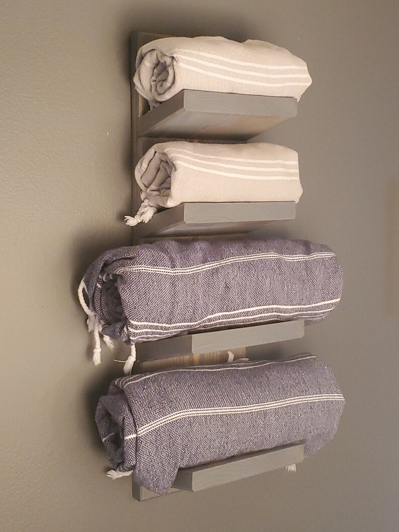 May include: A wooden wall-mounted towel rack with three shelves, each holding a rolled towel. The towels are beige and blue, with white stripes. The rack is painted gray and mounted on a gray wall.