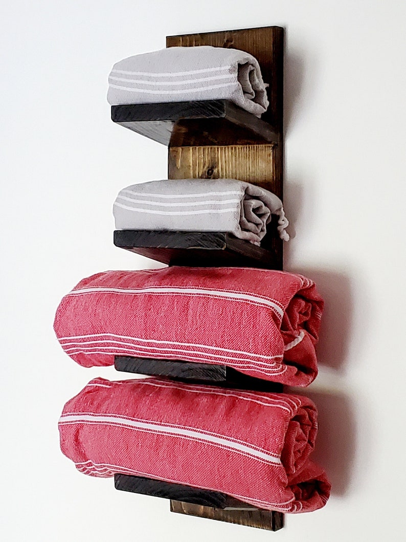 May include: A rustic wooden towel rack with three shelves. The shelves hold rolled towels in gray and red with white stripes. The wood has a dark stain, creating a contrast with the light-colored towels.