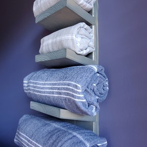 May include: A wooden towel rack with four shelves, painted a muted blue-green. The shelves hold rolled-up towels in shades of gray and blue, some with white stripes. The rack is mounted on a purple wall.