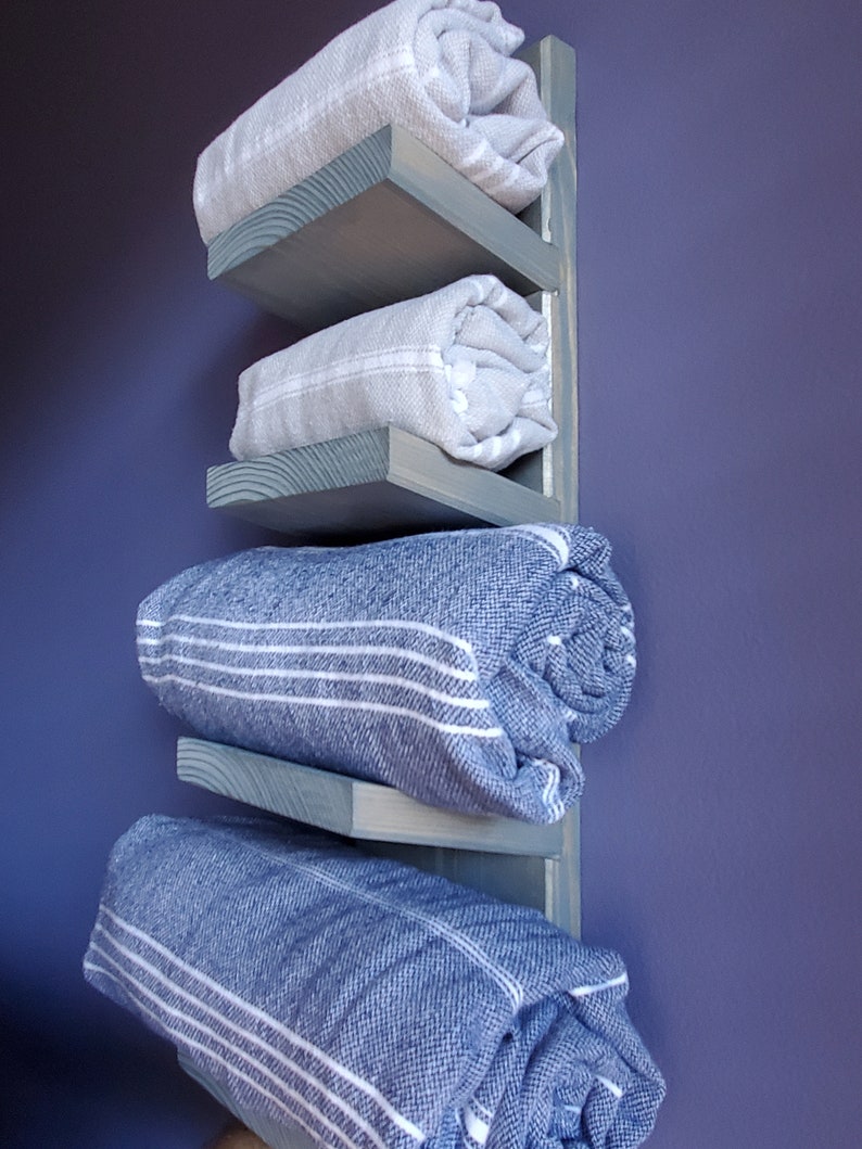 May include: A wooden towel rack with four shelves, painted a muted blue-green. The shelves hold rolled-up towels in shades of gray and blue, some with white stripes. The rack is mounted on a purple wall.