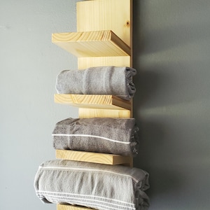 May include: A wooden wall-mounted towel rack with four shelves. The rack is made of light-colored wood and holds rolled gray towels. The towels have white stripes and fringe. The rack is mounted on a gray wall.