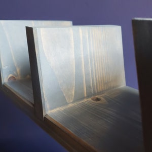 May include: A gray wooden floating shelf with three vertical supports. The shelf is made of stained wood and has a modern, minimalist design. The shelf is mounted on a purple wall.