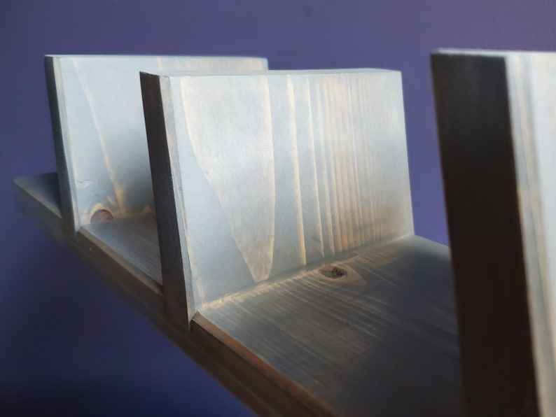 May include: A gray wooden floating shelf with three vertical supports. The shelf is made of stained wood and has a modern, minimalist design. The shelf is mounted on a purple wall.