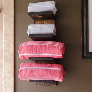 May include: A wall-mounted towel rack made of dark stained wood, holding four rolled towels. Two gray towels with white stripes are stacked above two red towels with white stripes. The rack is mounted on a brown wall.