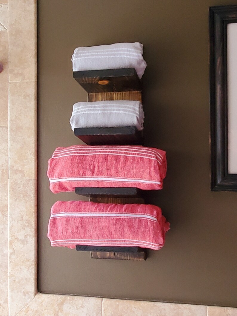 May include: A wall-mounted towel rack made of dark stained wood, holding four rolled towels. Two gray towels with white stripes are stacked above two red towels with white stripes. The rack is mounted on a brown wall.