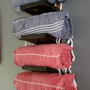 May include: A wall-mounted towel rack with four rolled towels. Two blue towels with white stripes are stacked above two red towels with white stripes. The towels are held by dark wooden shelves.