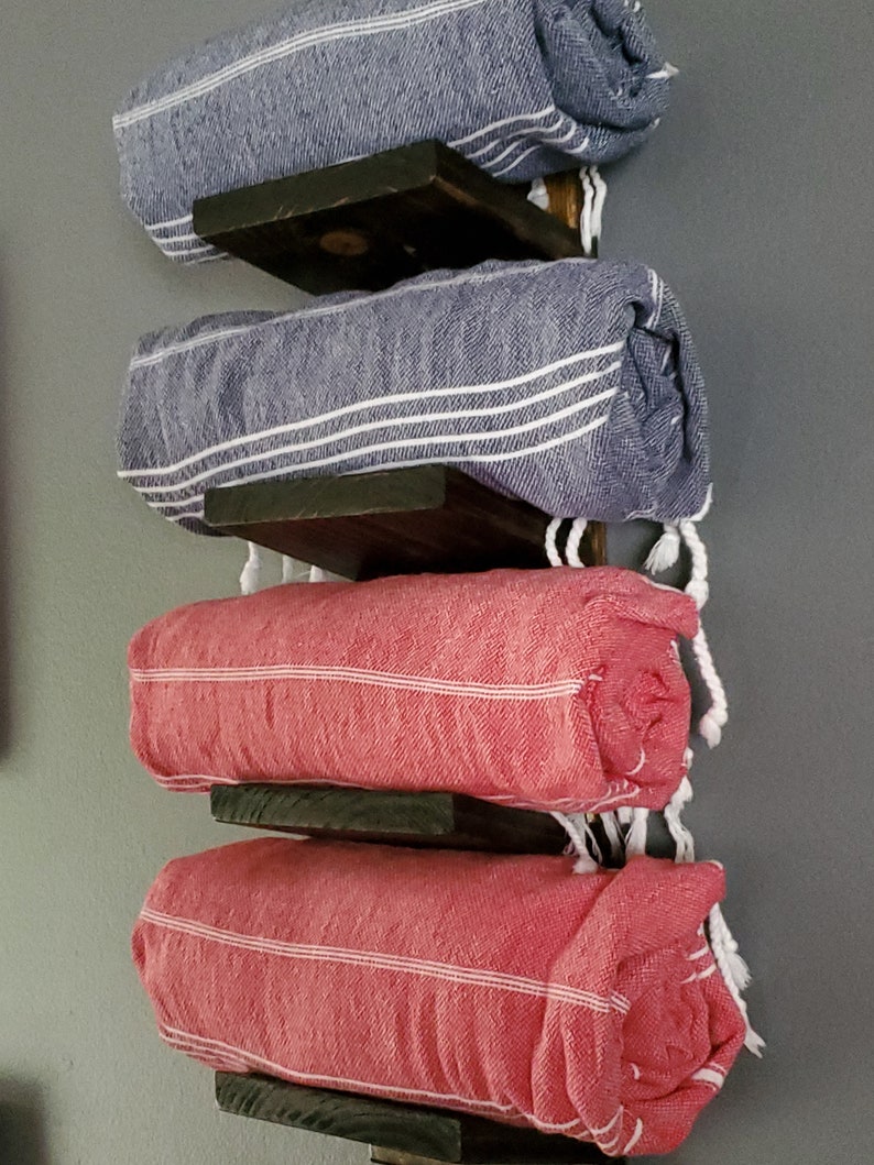 May include: A wall-mounted towel rack with four rolled towels. Two blue towels with white stripes are stacked above two red towels with white stripes. The towels are held by dark wooden shelves.