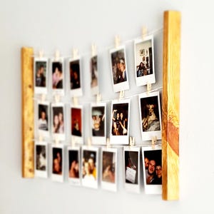 May include: A wooden photo display with multiple instant photos hanging from strings. The photos are held in place by small wooden clothespins. The wooden frame is a light brown color and is mounted on a light gray wall.