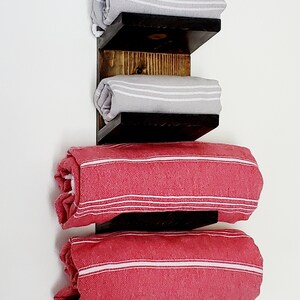 May include: A wooden towel rack with three shelves, holding rolled towels. The top two shelves display gray towels with white stripes, while the bottom two hold red towels with white stripes. The rack is made of dark stained wood.