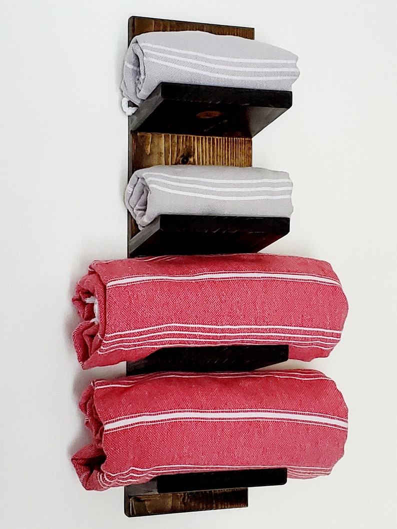 May include: A wooden towel rack with three shelves, holding rolled towels. The top two shelves display gray towels with white stripes, while the bottom two hold red towels with white stripes. The rack is made of dark stained wood.