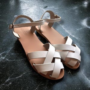 Op de afbeelding: Witte leren sandalen met een gekruist bandontwerp. De sandalen hebben een gespsluiting en een platte zool.