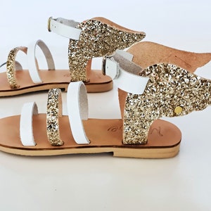 Peut inclure: Sandales en cuir blanc avec des lanières à paillettes dorées et des ailes à paillettes dorées. Les sandales ont une fermeture à boucle et une semelle plate.