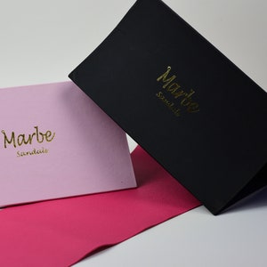 May include: Two boxes with the text "Marbe Sandals" in gold lettering. One box is black and the other is pink. Both boxes are sitting on a pink surface.