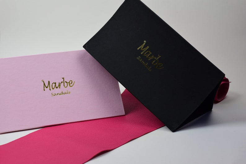 May include: Two boxes with the text "Marbe Sandals" in gold lettering. One box is black and the other is pink. Both boxes are sitting on a pink surface.
