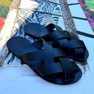 Puede incluir: Un par de sandalias de cuero negro con un diseño tejido. Las sandalias tienen una suela simple y plana y el nombre de la marca "Nanné" está estampado en las tiras.