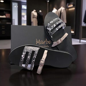 May include: Black sandals with a snakeskin pattern on the straps and a buckle fastening. The sandals are displayed with a black box that has the brand name "Marbe sandals" in gold lettering.