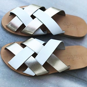 May include: White and gold leather sandals with a woven design. The sandals have a brown leather sole and the brand name "Jolipa" is printed on the sole.