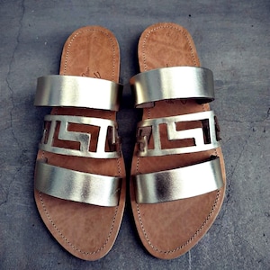 May include: A pair of gold leather sandals with three straps and a cut-out design. The sandals have a brown leather sole.