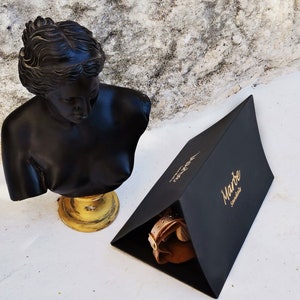 Pu&ograve; includere: Una scatola triangolare nera con lettere dorate che recitano "Marte Leather" &egrave; appoggiata su una superficie bianca. Un busto nero di una donna &egrave; appoggiato su un piedistallo dorato a sinistra della scatola.