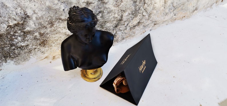 Pu&ograve; includere: Una scatola triangolare nera con lettere dorate che recitano "Marte Leather" &egrave; appoggiata su una superficie bianca. Un busto nero di una donna &egrave; appoggiato su un piedistallo dorato a sinistra della scatola.