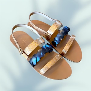 May include: A pair of brown leather sandals with gold and white detailing. The sandals have a gold textured pattern on the straps and a braided blue strap. They have a buckle fastening and a flat sole. The sandals are suitable for summer wear.